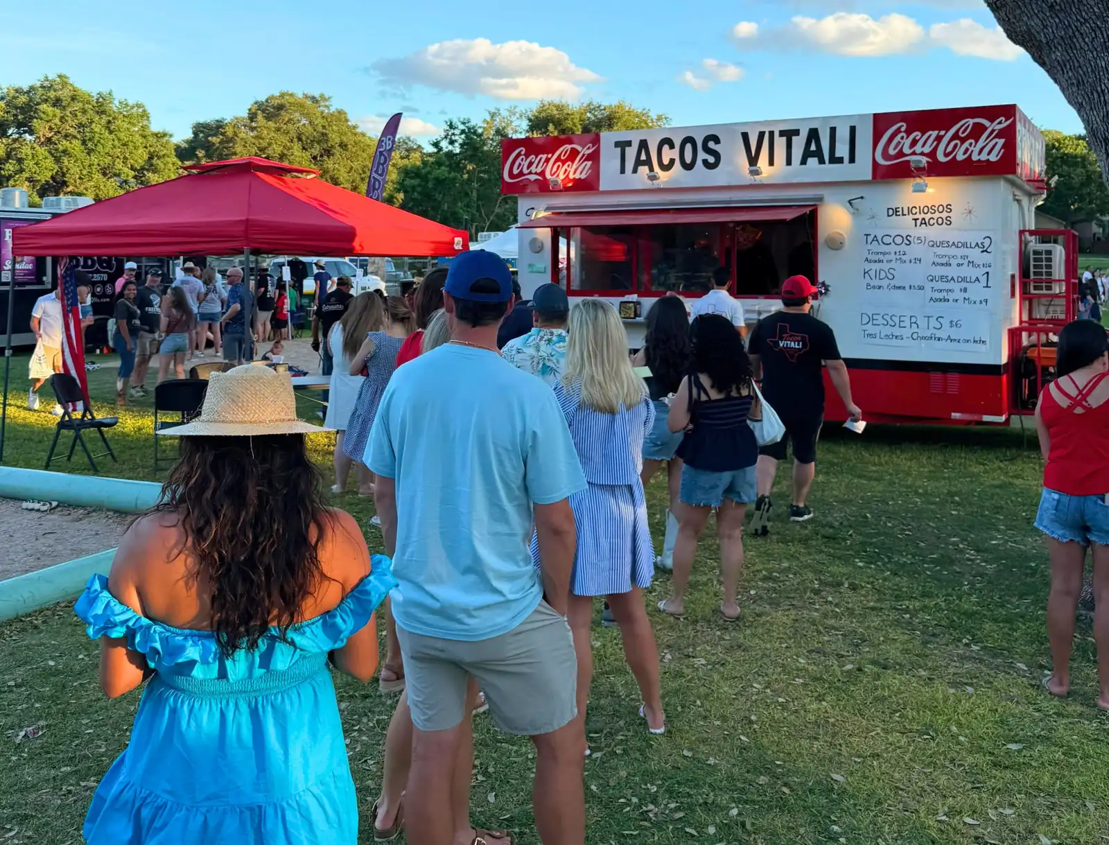 A Tacos Vitali trailer serving a line of people at a park